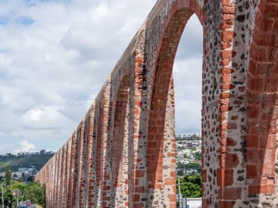 A long stone archway with a red brick wall. The archway is on a street with cars driving by. Historic center of Querétaro, colonial architecture, decorations for the celebration of Mexico's Independence Day.